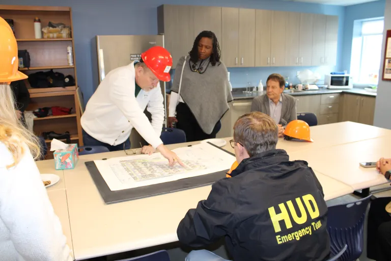 Dan McColley, Executive Director of Flatirons Habitat for Humanity, leans over a conference table to point out details on a large printed site map, while several others look on in a break room setting.