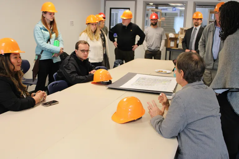 A group of visitors wearing orange hard hats gathers around a conference table during a briefing at BoulderMOD.