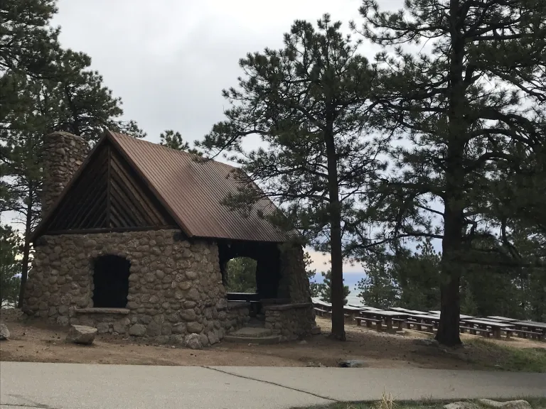 The Stone Shelter surrounded by pine trees with picnic tables in the background.