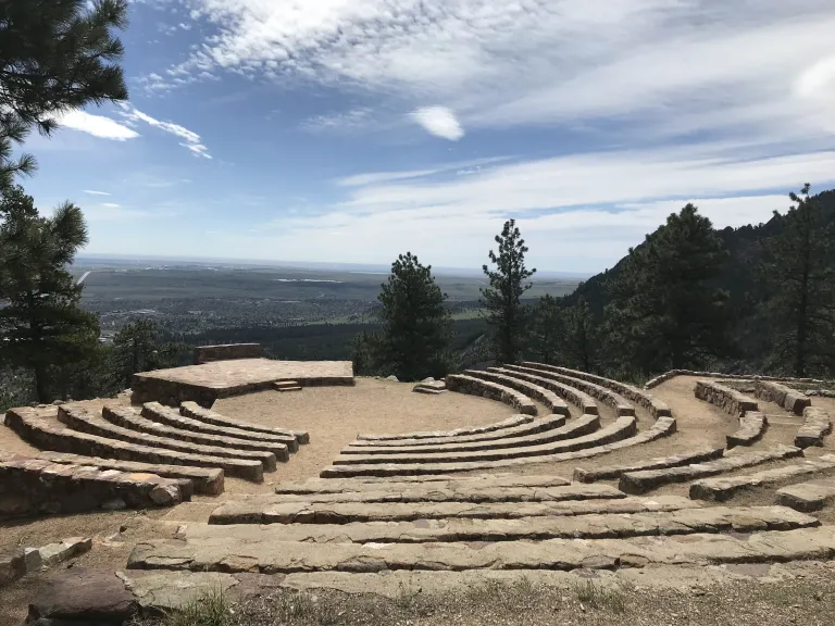 Rows of stone seats arc around the center circle and stage of the amphitheater, with the Flatirons visible to the right.