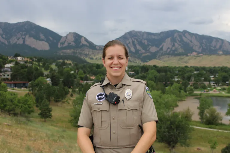 Urban Ranger Caitlyn Imfeld stands above Harlow Platts Park, with the Boulder Flatirons and Viele Lake visible in the background. 