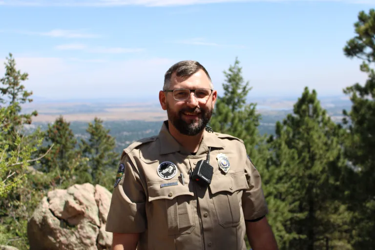 Urban Ranger Brian Bauer stands on top of Flagstaff Mountain, framed by trees and blue skies, with a view of Boulder in the background. 