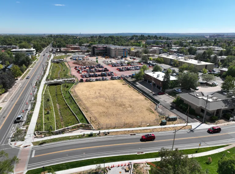 Aerial view of the flood channel along the roadway at left, next to a cleared dirt lot, a parking area, and a multistory building under construction.
