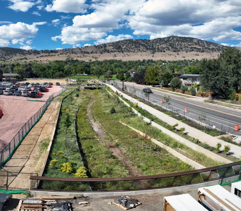 Grassy Alpine-Balsam flood channel with concrete walls, seen from a bridge railing, with a sidewalk and road on the right, a gravel parking area on the left, and foothills beyond.