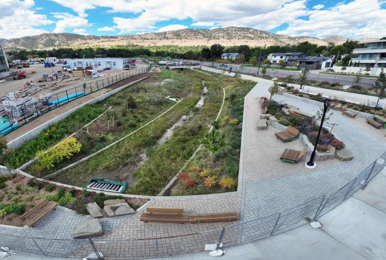 Curving flood channel with native grasses, wildflowers, and a thin stream of water, beside a brick-paver plaza with wooden benches, boulders, and flowering plantings.