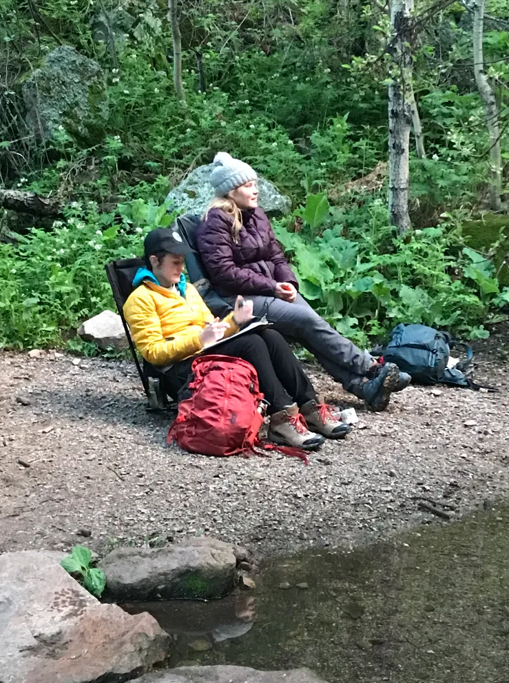 Two people sitting outside next to a body of water writing on a clipboard.