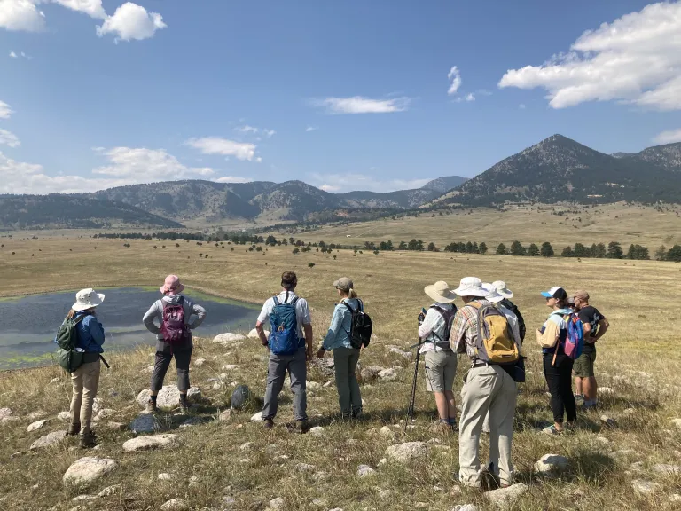 Several people standing in a rocky prairie looking out at a small pond.