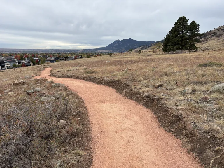 A smooth dirt trail winds downhill with a few of the Flatirons in the distance. 