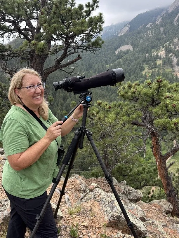 A woman standing on a mountain side with a very large lens camera on a tripod.