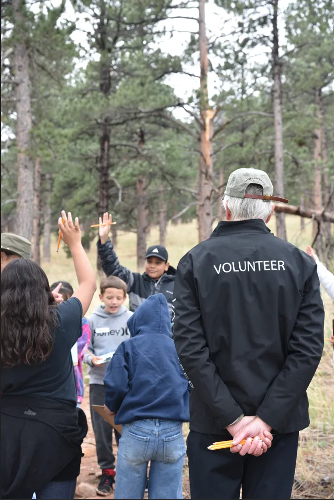 The back of a person with a jacket that reads 'Volunteer'. He is looking a bunch of kids with their hands raised.