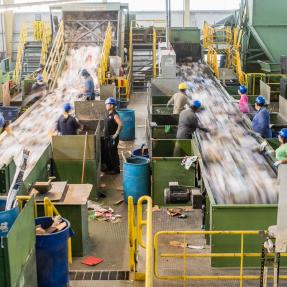 Image of a recycling center, two parallel rows of sorting machines  with workers on eitehr side. 