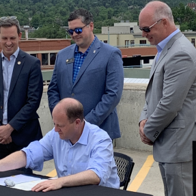 Governor Polis, seated, signs a bill on a Boulder rooftop, surrounded by Colorado state legislators. 