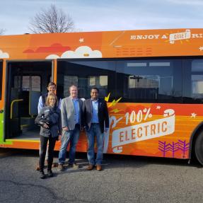 Government officials stand in front of an orange, all-electric city bus