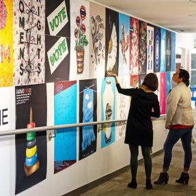 Two people look at an exhibition of large posters of graphic art on a wall,