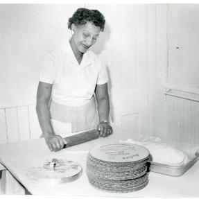 A woman rolling out pie dough besides a stack of pie crusts.