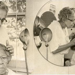 A woman wearing black-rimmed glasses adjusts her sisters hair at a salon. The two can be seen reflected in the mirror. 