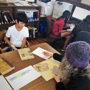 Middle schoolers and an adult look at old newspaper clippings and worksheet papers. 