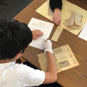 A middle schooler looks at an old newspaper clipping and writes notes into a worksheet. He is wearing a white shirt and a white latex glove while he writes.