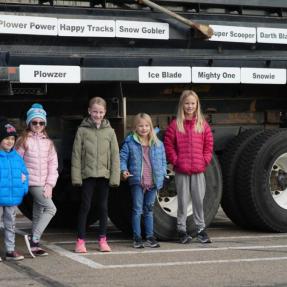 six children standing besides a snowplow with plaques of snowplow names