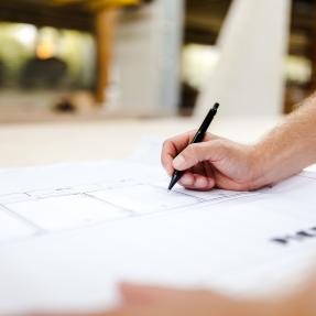 Image of a hand holding a pen drawing a site plan. Blurred background appears to be a constuction site.