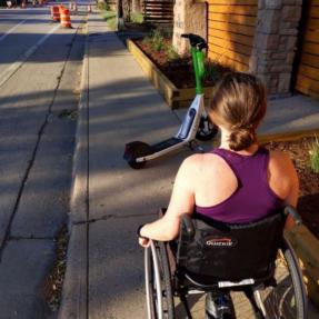 Person on wheelchair traveling on sidewalk blocked by scooter