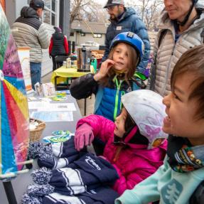 Kids in colorful jackets and helmets outside watch a spinning prize wheel at a table for Winter Bike to Work Day