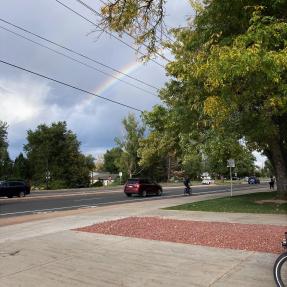 A rainbow near a road. There is a sidewalk and a large driveway that leads to it.