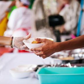 Volunteer wearing a plastic glove passing a bowl of food to someone. Only hands and arms are visible.