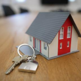 House key on a keychain with a metal house charm in front of a red and white model home.