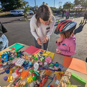 City staff speaks with a child next to a table of miscellaneous toys for a transportation activity.