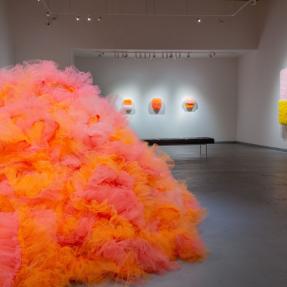 Orange and pink tulle fabric sewn together into an abstract sculpture on the floor of an art gallery.