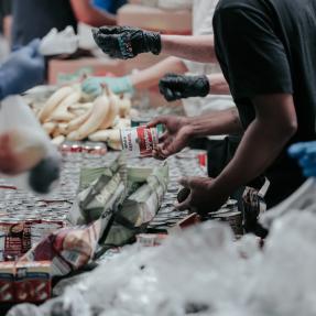 Scene from a food bank, picture shows the hands of volunteers over a table full of various food items. 