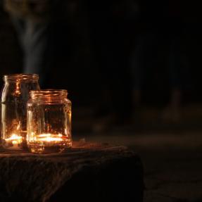 A dark scene with two glass jars lit up with tea light candles sitting on a tree stump 