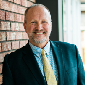 A headshot of Dave Ruppel. In the photo, Ruppel is a middle-aged white man with graying hair and a beard. He is smiling at the camera and wearing a suit with a yellow tie and blue dress shirt.