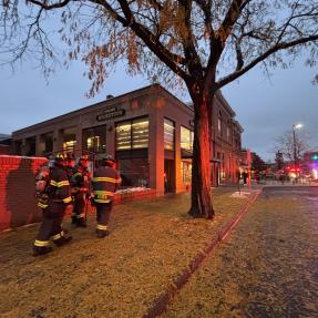 Four Boulder firefighters dressed in full gear approaching the Boulder Bookstore at dusk lit up by the blue and red lights from nearby vehicles. 