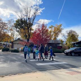 A group of 6 students and 2 parents walking across the crosswalk on Manhattan Drive near Iroquois Drive. A car is stopped at the stop sign, yielding to the group of pedestrians.