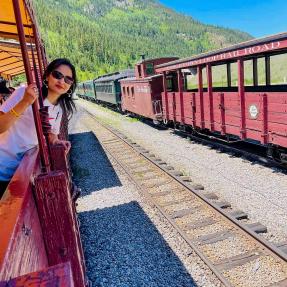 Woman leans head out of a train car. There is another train in the background and there are mountains in the background behind the trains. 