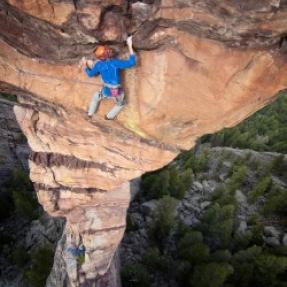 Vertigo-inducing view from above of a climber on a thin rock formation with trees far below.