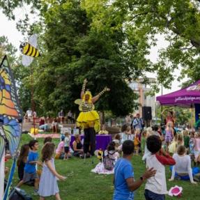 Outdoor festival scene at a park with children and families watching a performer in a bright yellow bee costume on stilts. A purple canopy with the Jeff and Paige logo is set up near a large tree, and colorful butterfly and bee decorations are visible among the crowd.