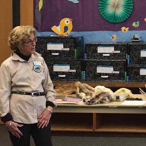 Woman in khaki shirt standing in a colorful classroom surrounded by animal pelts.
