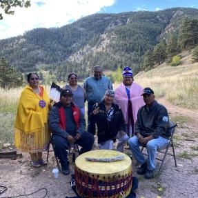 Tribal elders seated around a large drum with the Boulder mountains in the background.