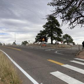 Route à double sens avec une grande aire de stationnement bloquée par une barrière métallique.