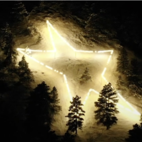 Large star made from lights on the side of a snowy mountain with pine tree around it.