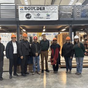 A delegation from the Japan Off-site Construction Association, Councilmember Taishya Adams and Habitat and city staff pose in front of the BoulderMOD sign on the factory floor.