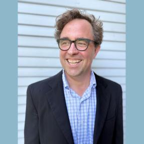 Headshot of Blythe Bailey, a white man with brown hair, smiling in a black suit jacket, blue plaid shirt and glasses