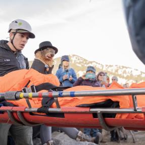 People wearing helmets carry a stuffed marmot on a bright orange stretcher