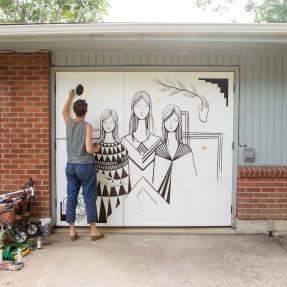 Artist painting a mural of women on the garage door of a house