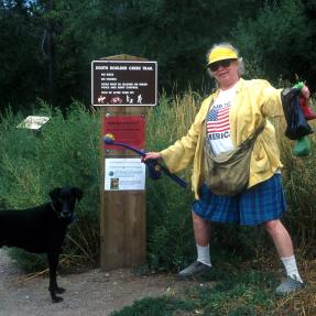 Woman carrying two poop bags to a trash can while holding her dog on leash.