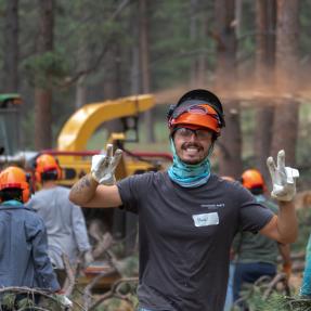 Volunteer wearing helmet and safety gear smiles and poses in front of group working in a forest with a chipper.