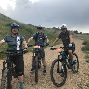 Three bikers on a wide gravel trail, wearing helmets and smiling, two of their bikes have orange placards that read "Boulder Mountain Bike Patrol".